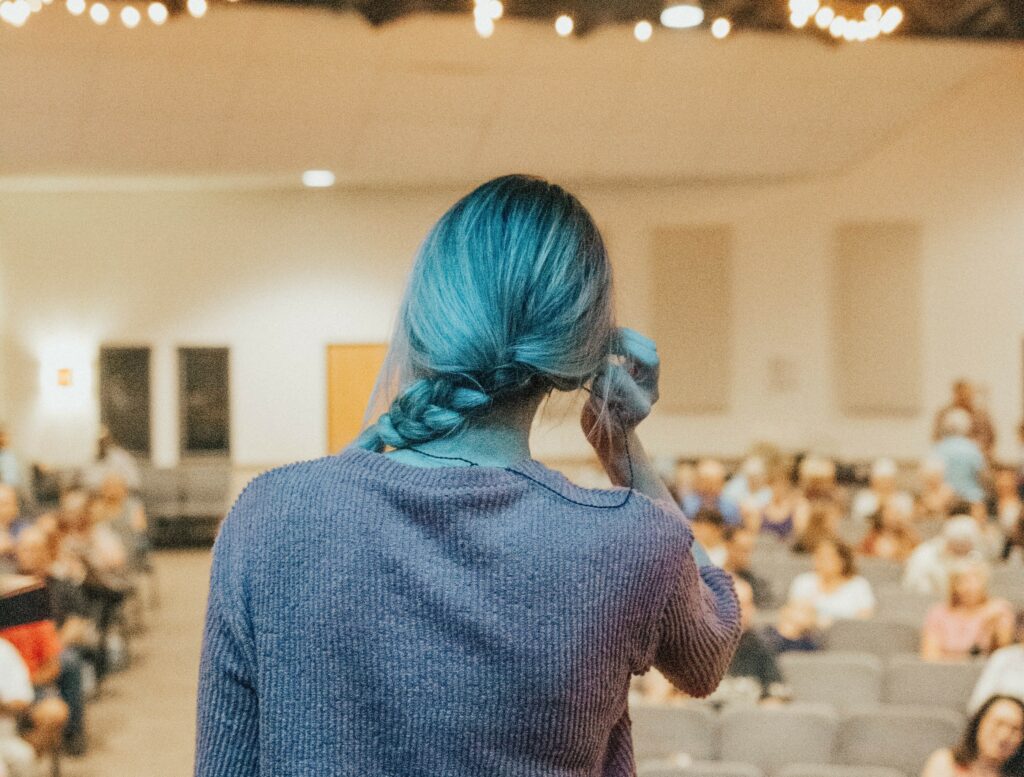Une femme, de dos, devant une salle comble, sa posture montre qu'elle manque de confiance en elle